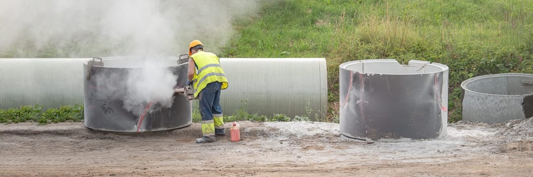 Road reconstruction process. A worker installs a rainwater collector. A man in a helmet and a respirator saws a hole in a concrete pipe with a saw. The concept of reconstruction and construction