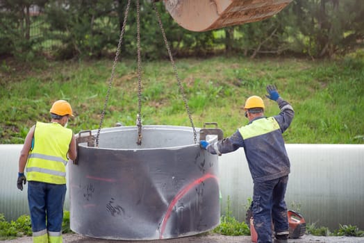 Workers are working on the construction site. An excavator uses a chain to lift a concrete pipe to install a rainwater or sewer collector. Reconstruction of the road and communications