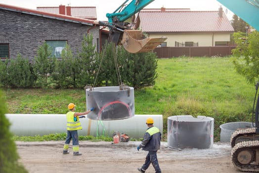 Workers are working on the construction site. An excavator uses a chain to lift a concrete pipe to install a rainwater or sewer collector. Reconstruction of the road and communications