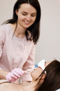 Beautician holds a wooden spatula and applies a transparent gel with anesthetic to the woman's face and prepares the skin for the laser hair removal procedure. Laser mustache hair removal.