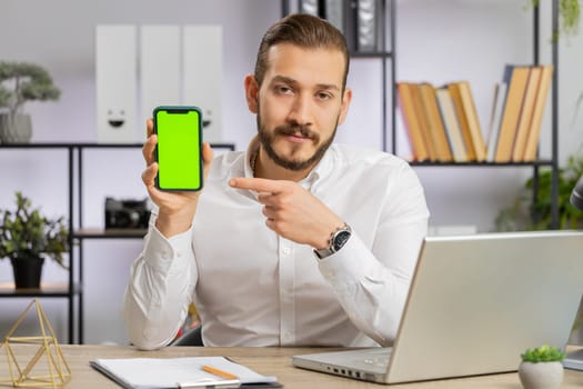 Middle eastern developer business man holding pointing on smartphone with green screen chroma key mock up recommend good application promotional sale offer. Adult freelancer guy at office workplace