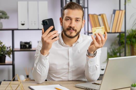 Young middle eastern boss business man using credit bank card and smartphone while transferring money, purchases online shopping ordering delivery. Freelancer guy at office workplace looking at camera