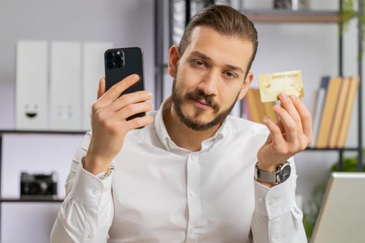 Young middle eastern boss business man using credit bank card and smartphone while transferring money, purchases online shopping ordering delivery. Freelancer guy at office workplace looking at camera