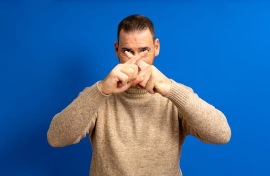 Hispanic man in his 40s crossing his fingers in stop and protest against the total oppression of the elites towards the proletariat, isolated on blue background