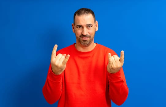 Bearded hispanic man in his 40s wearing a red sweatshirt making a rock gesture in concept of rebellion and illegality, isolated over blue background.