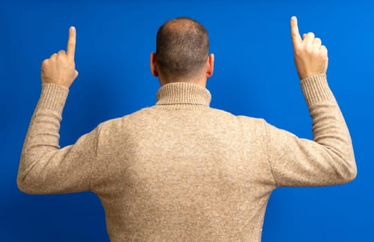 Portrait of a young man from behind, raising both hands and pointing the index finger up, wearing a beige pullover, isolated on a blue background.