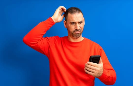 Bearded hispanic man in his 40s wearing a red sweatshirt with an expression of disbelief while looking at a mobile and scratching his head not understanding the concept, isolated over blue background