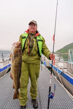 Male fisherman holding a huge fish Cod. Norway Fishing tourism. Senior fisherman in ocean, fjord fishing. Fishing in Lofoten, Senja, Alta, Tromso.