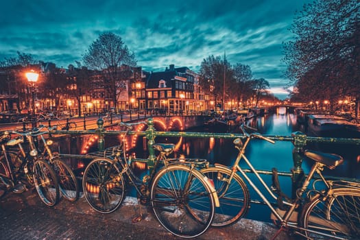 Night view of Amterdam cityscape with canal, bridge with bicycles and medieval houses in the evening twilight illuminated. Amsterdam, Netherlands