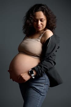 Curly haired Latina pregnant woman, expectant mother in beige bra and blue jeans, touching her big belly in the ninth month of her pregnancy, smiling, isolated over fashion gray studio background.
