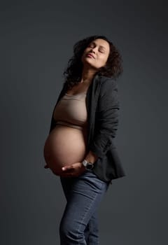 Studio portrait of a multi-ethnic woman 30s, delightful gorgeous pregnant expectant mother holding her belly, posing with her eyes closed over isolated gray background. Beautiful pregnancy. Maternity