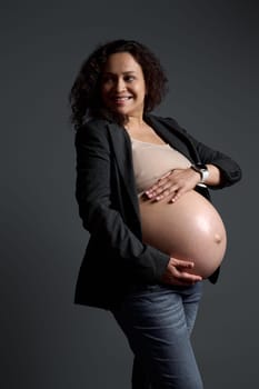 Attractive Latin American pregnant woman, happy smiling mother expecting her baby, holding her big belly in the week 36 of carefree pregnancy, looking aside a copy ad space, isolated gray background