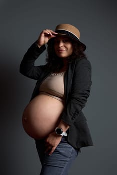 Attractive feminine pregnant woman, mother-to-be, wearing stylish blazer over lingerie and jeans, smiling looking at camera, posing bare belly on gray background. The concept of beautiful pregnancy