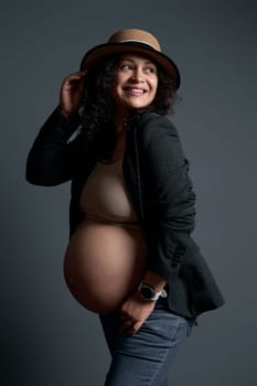 Stunning pregnant woman in straw hat and stylish gray blazer over lingerie, smiling with a beautiful toothy smile, looking aside a copy ad space on studio background. Happy carefree healthy pregnancy