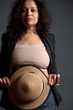 Close-up authentic portrait of a multi-ethnic confident adult pregnant woman in stylish blazer, looking at camera, holding a straw hat, hiding her belly, isolated on fashion gray studio background