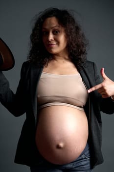 Multiethnic pregnant woman, gravid expectant mother pointing finger at her big pregnant belly in the ninth month of carefree happy pregnancy, smiling, looking at the camera, isolated studio background