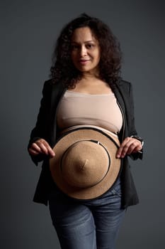 Pregnant woman in a gray blazer and blue denim, smiling at camera, hiding her big belly with a straw hat, isolated over gray studio background. Beautiful pregnancy. Women's health. Gynecology concept