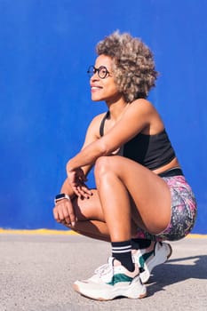 vertical portrait of a smiling african american sportswoman squatting in a blue background, concept of sport and healthy lifestyle