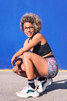 vertical portrait of an african american sportswoman squatting looking at camera smiling happy in a blue background, concept of sport and healthy lifestyle