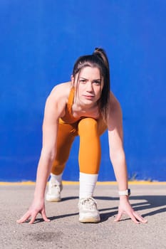 vertical portrait of a focused sportswoman in position to start a race with orange sportswear in a blue background, concept of sport and active lifestyle, copy space for text