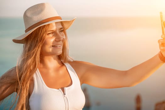 Selfie woman in a hat, white tank top, and shorts captures a selfie shot with her mobile phone against the backdrop of a serene beach and blue sea