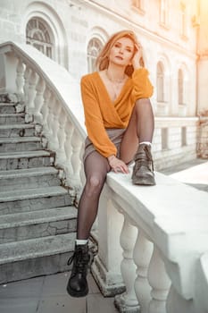 A middle-aged woman looks like a good blonde with curly beautiful hair and makeup on the background of the building. She is wearing a yellow sweater