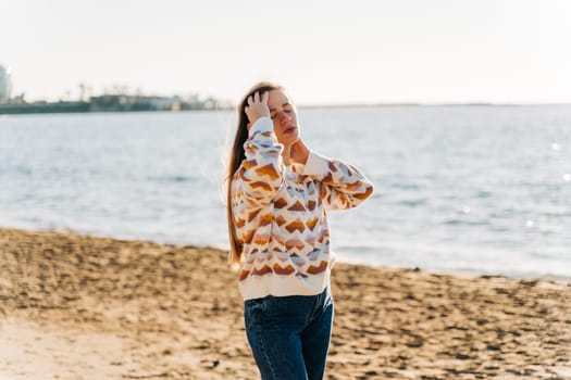 Young beautiful woman in cozy sweater smiling and enjoying sunbathe on the sand beach near winter ocean. Cute attractive girl relaxing under the sun on autumn seaside shore.