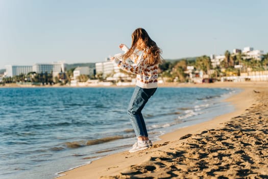 Side portrait of young beautiful woman in cozy sweater running to the winter sea waves. Cute attractive girl having fun jumping near autumn ocean on the beach sand.