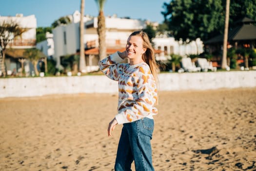 Young beautiful woman in cozy sweater smiling enjoying winter sunbathe on the sand beach with white beach bungalows in the background. Cute attractive girl relaxing under sun on autumn seaside shore.