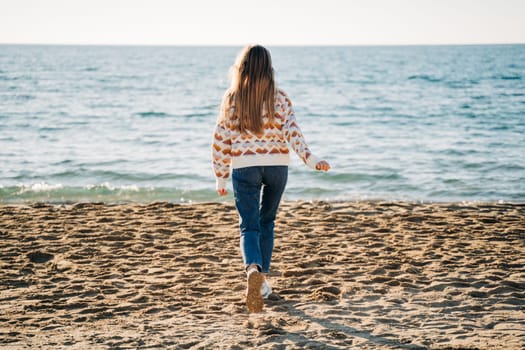 Back portrait of a girl in cozy sweater and jeans running on the winter beach to the sea. Woman walking on the autumn seashore sand and enjoying the view of a fall ocean waves.