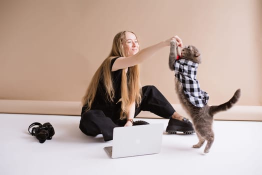 A blogger freelancer in a black is working on a laptop apple and playing, eating with Scottish straight cat. blonde assistant in video production with computer MacBook and camera Lumix GH5