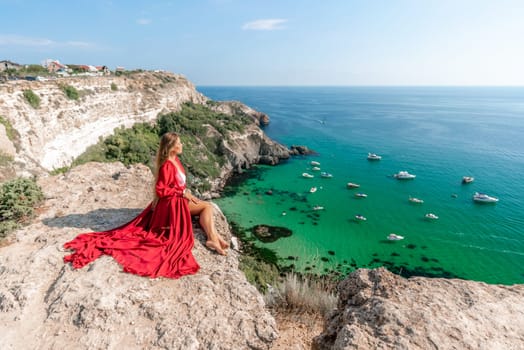 Woman red dress sea. Happy woman in a red dress and white bikini sitting on a rocky outcrop, gazing out at the sea with boats and yachts in the background