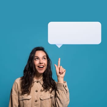 Glad surprised millennial european brunette woman in casual got idea, showing finger up on blank white communication balloon, isolated on blue background, studio. Shocking thought, exciting idea