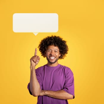 Your opinion matters concept. Cheerful handsome millennial black guy in purple t-shirt pointing index finger up at white blank communication bubble, smiling on yellow background, having nice idea
