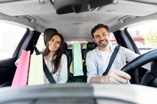 Cheerful beautiful millennial spouses driving home after shopping in mall, smiling. Happy man and woman sitting inside car, holding colorful shopping bags purchases. Black friday, season sale