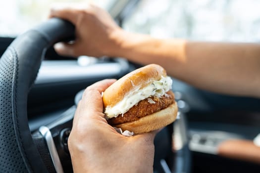 Asian lady holding hamburger to eat in car, dangerous and risk an accident.