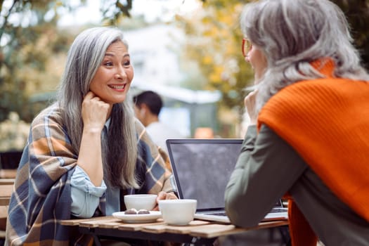 Positive mature Asian lady and grey haired friend with laptop sit together at table on outdoors cafe terrace cafe on nice autumn day