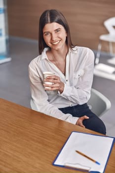 Joyful caucasian lady is drinking a coffee from a white paper cup while sitting on reception