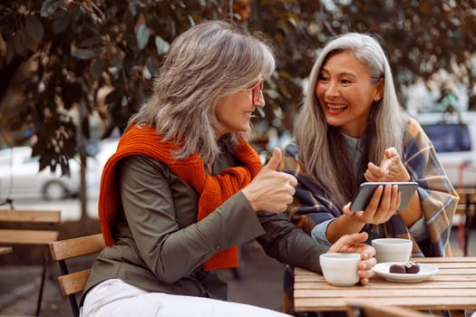 Cheerful senior Asian lady shows phone to grey haired friend at small table with coffee and candies in street cafe on nice autumn day