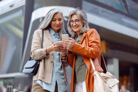 Positive grey haired mature ladies look at modern mobile phones on modern city street on nice autumn day. Old friends meeting