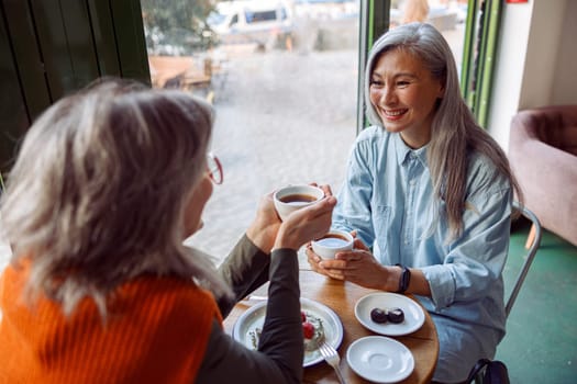Pretty happy mature women companions with natural grey hair hold cups of hot drinks sitting at small table in cozy cafe. Long-time friendship