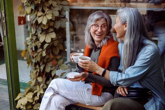 Positive senior Asian woman with friend clink cups of delicious drinks sitting on bench near cozy cafe outdoors. Long-time friendship