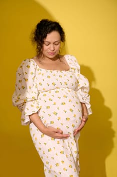 Multi-ethnic curly haired middle aged pregnant woman, gravid expectant mother in stylish patterned summer sundress, touching her big belly, isolated over yellow studio background. Beautiful pregnancy