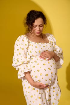 Multiethnic curly haired middle aged beautiful pregnant woman putting hands on her belly, enjoying baby kicks, isolated over yellow background. Pregnancy and maternity. Human fertility. Women's health