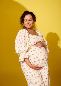 Authentic portrait of a beautiful pregnant woman in white stylish summer sundress, touching her big belly, smiling looking aside, isolated yellow studio background. Studio shot with light shadow