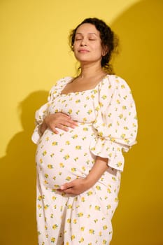 Happy pregnant woman, beautiful expectant mother putting hands on her belly, enjoying baby kicks, isolated over yellow background. Pregnancy and maternity. Human fertility. Women's health. Gynecology