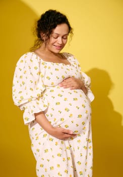 Multi-ethnic curly haired middle aged pregnant woman, gravid expectant mother in stylish patterned summer sundress, stroking her big belly, isolated over yellow studio background. Beautiful pregnancy