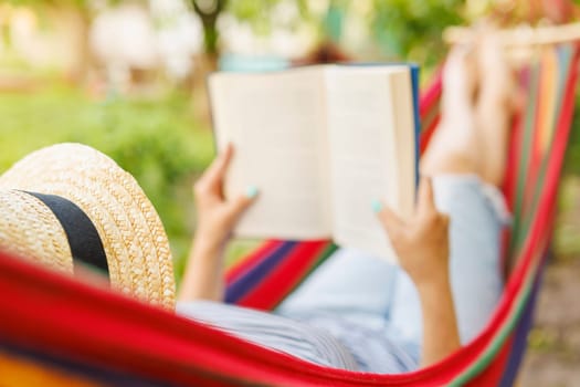 Young woman reading book while lying in comfortable hammock at green garden.
