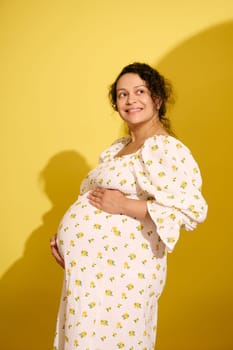 Studio portrait of a beautiful multi-ethnic pregnant curly haired woman, dressed in white summer dress, cutely smiling looking away, holding her belly, isolated over yellow studio background.