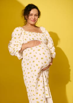 Beautiful curly haired pregnant woman in stylish summer sundress, gently caresses her belly, smiles looking at camera, isolated on yellow background. Beautiful pregnancy and maternity leave concept